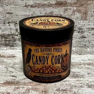 May include: A cylindrical black metal tin featuring a vintage-style label with the text The Ravens Finest Candy Corn. The label displays a black raven, a jack-o'-lantern, and a pile of candy corn. The lid also features matching graphics.