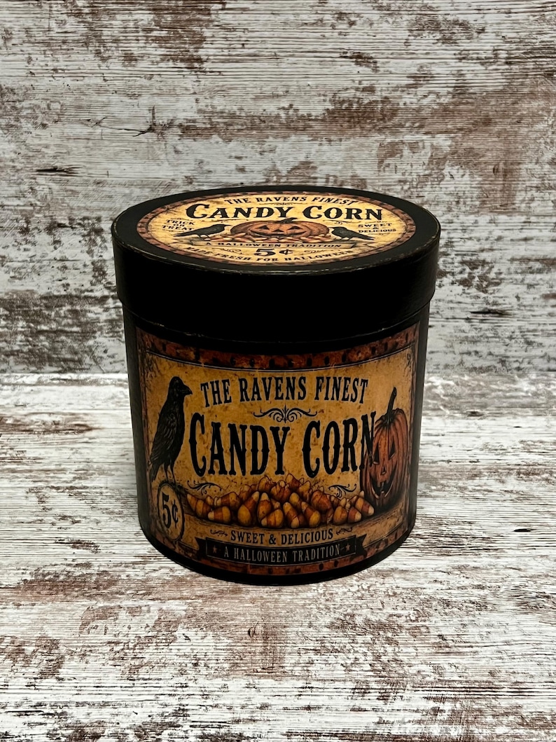 May include: A cylindrical black metal tin featuring a vintage-style label with the text The Ravens Finest Candy Corn. The label displays a black raven, a jack-o'-lantern, and a pile of candy corn. The lid also features matching graphics.