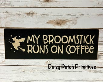 May include: A black wooden sign with the phrase "MY BROOMSTICK RUNS ON COFFEE" in white lettering. A witch on a broom is depicted on the left side. The sign is a Halloween or fall-themed decorative item.