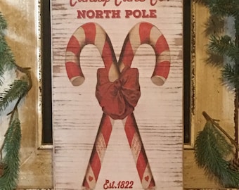May include: A rectangular wooden sign with a distressed white background features two crossed red and white striped candy canes tied with a red bow. The sign reads "Candy Cane Co. North Pole" and "Est. 1822 Peppermint Candy Canes."