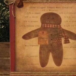 May include: A framed print featuring a gingerbread man design with a red and brown color scheme. The print includes a recipe for "Olde Ginger Bread Man Cookies" and is decorated with a jute bow and bell.