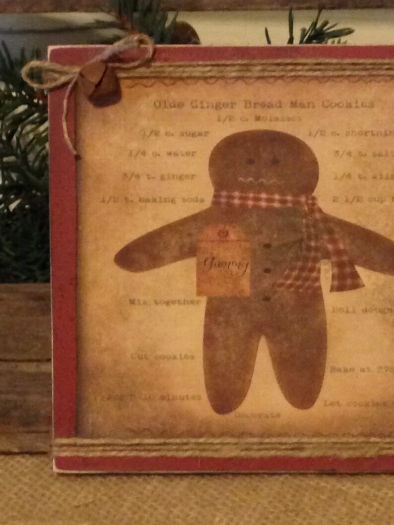 May include: A framed print featuring a gingerbread man design with a red and brown color scheme. The print includes a recipe for "Olde Ginger Bread Man Cookies" and is decorated with a jute bow and bell.
