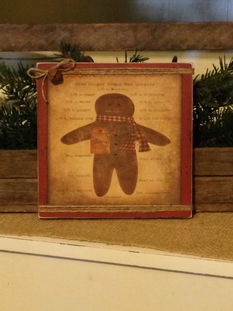 May include: A rustic, framed sign featuring a gingerbread man illustration. The sign has a red border and a twine accent. The gingerbread man wears a plaid scarf and holds a tag that says "Joy". The sign also includes a recipe for "Olde Ginger Bread Man Cookies".