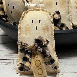May include: Handmade ghost-shaped fabric ornaments with black button eyes and a frayed edge. Each ghost has a black and tan plaid bow and a tag that says "BOO". The ornaments are part of a Halloween or fall decor collection.
