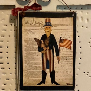 May include: A wooden sign with a black frame featuring a cartoon illustration of a man in a blue jacket and red and white striped pants holding a bald eagle and an American flag. The sign has the text "We the People" and the text of the US Constitution printed on it.