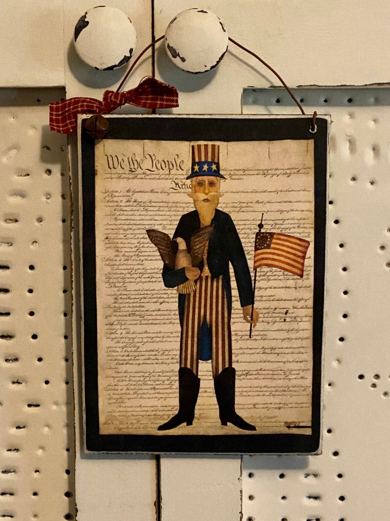 May include: A wooden sign with a black frame featuring a cartoon illustration of a man in a blue jacket and red and white striped pants holding a bald eagle and an American flag. The sign has the text "We the People" and the text of the US Constitution printed on it.