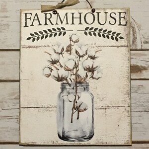 May include: A white wooden sign with the text "FARMHOUSE" in black lettering. The sign features a painted image of a glass jar filled with cotton bolls.