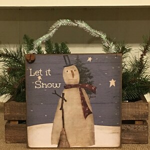 May include: Wooden sign with a snowman illustration and the text "Let it Snow." The snowman wears a top hat and a scarf. The sign has a silver tinsel hanger and a small bell. The background is a blue and white winter scene.