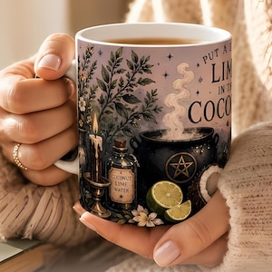 May include: A white ceramic mug featuring a colorful illustration of a black cauldron with a pentagram, a lit candle, a bottle labeled Coconut Lime Water, lime slices, and text that reads Put a Lime in the Coconut.