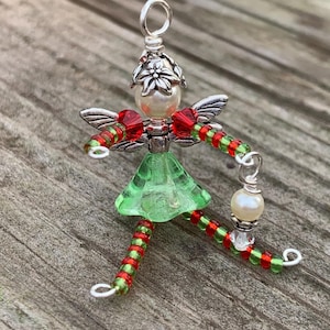 May include: A handmade fairy ornament with a silver headpiece and wings. The fairy has a green skirt, red and green beaded legs, and a pearl-like bead. The ornament is on a wooden surface.