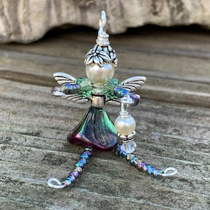 May include: A decorative figurine crafted from wire, beads, and crystals. The figure features a white pearl head with a silver flower cap, iridescent green and purple glass wings, a dark purple skirt, and long legs made of multi-colored metallic beads.