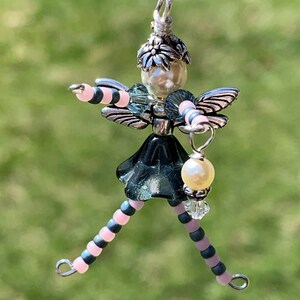 Könnte beinhalten: Ein Perlen-Feen-Anhänger mit rosa-schwarzen gestreiften Beinen, einem grünen Glasrock und silbernen Flügeln. Die Fee hält eine Perle und eine Kristallperle.