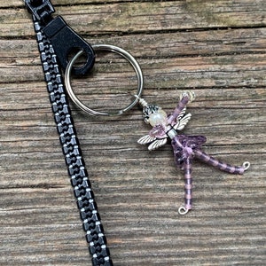 May include: A purple beaded fairy charm with silver wings and a pearl bead on a silver key ring attached to a black zipper pull.