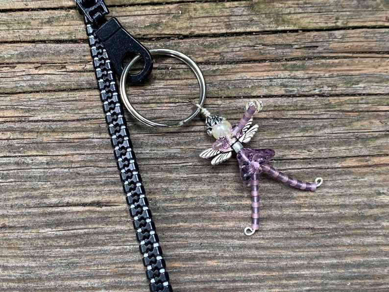 May include: A purple beaded fairy charm with silver wings and a pearl bead on a silver key ring attached to a black zipper pull.