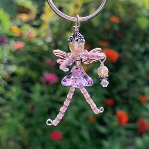 May include: A purple beaded fairy charm with wings, a pearl crown, and a pearl and crystal dangle. The charm is attached to a silver key ring.