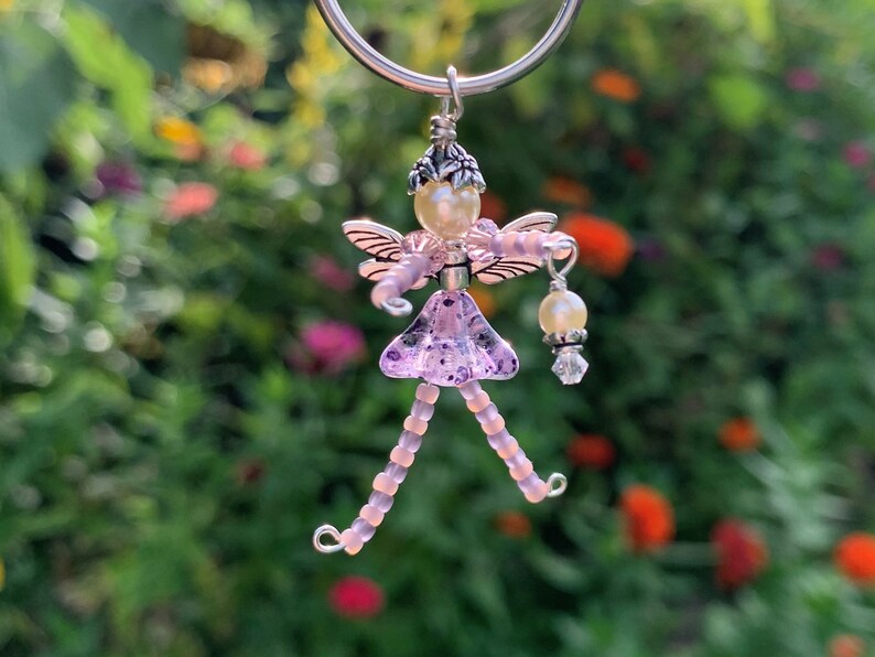 May include: A purple beaded fairy charm with wings, a pearl crown, and a pearl and crystal dangle. The charm is attached to a silver key ring.