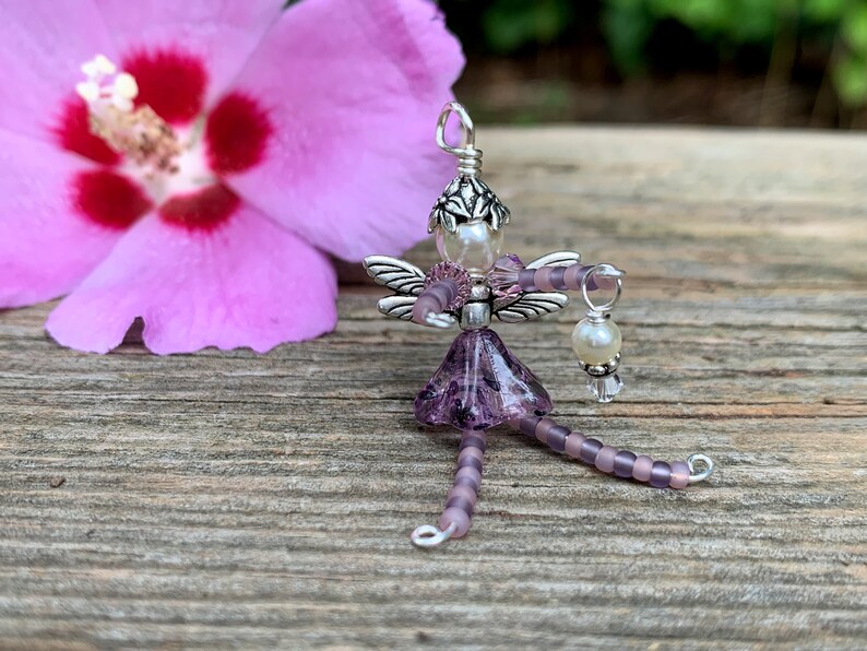 May include: A purple beaded fairy charm with silver wings and a pearl accent. The fairy is holding a small pearl and crystal charm.