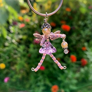 May include: A pink beaded fairy charm with silver wings and a pearl head. The fairy is holding a pearl and crystal charm. The charm is hanging from a silver ring.