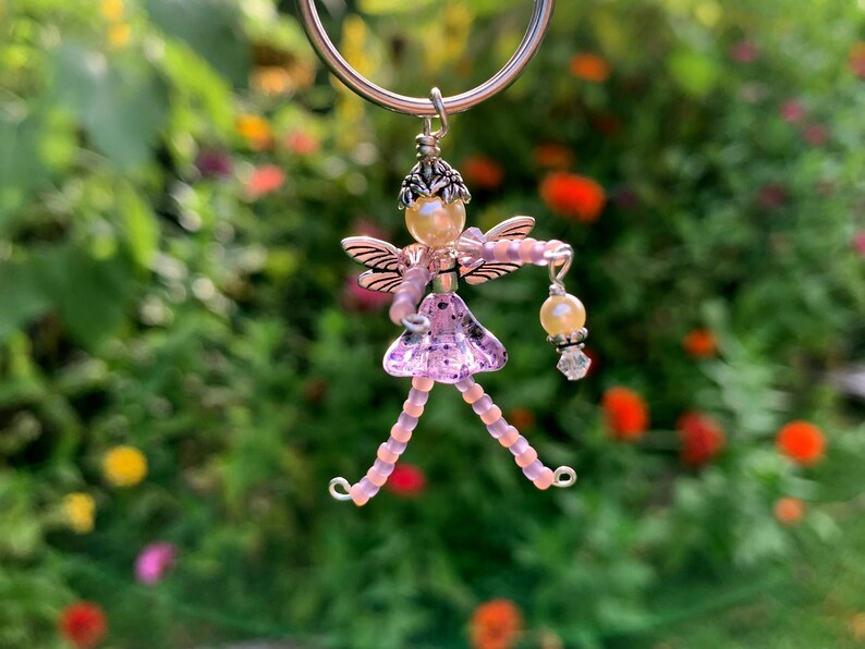 May include: A pink beaded fairy charm with silver wings and a pearl head. The fairy is holding a pearl and crystal charm. The charm is hanging from a silver ring.
