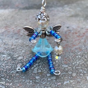 May include: A handcrafted fairy figurine with a pearl head, silver wings, and a light blue dress. The fairy holds a small lantern and has beaded legs and arms. The fairy's head is adorned with a silver floral crown.