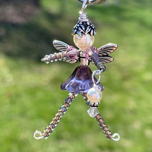 May include: A beaded fairy charm with iridescent purple wings, a pearl head, and a silver flower crown. The fairy is holding a pearl and a crystal bead.