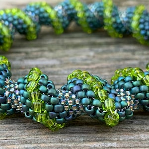 Puede incluir: Un primer plano de un brazalete de cuentas verdes y plateadas. El brazalete está hecho de pequeñas cuentas redondas que están dispuestas en un patrón en espiral. Las cuentas son una mezcla de colores verdes, plateados e iridiscentes.