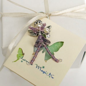 May include: A purple and silver fairy charm with beaded wings and a beaded dress. The charm is on a white card with the text "Love Magick" printed on it.