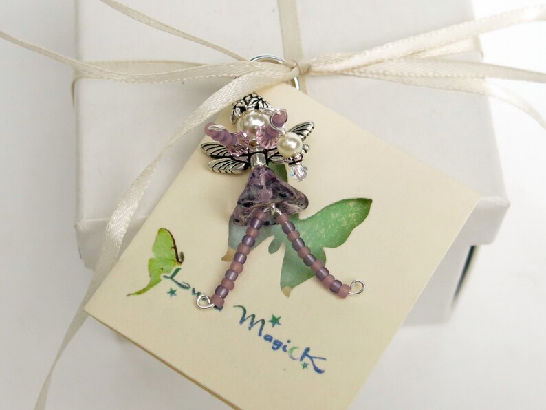 May include: A purple and silver fairy charm with beaded wings and a beaded dress. The charm is on a white card with the text "Love Magick" printed on it.