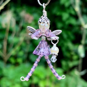 May include: A beaded fairy charm with a purple dress, wings, and a pearl and crystal embellishment. The fairy is holding a small lantern.