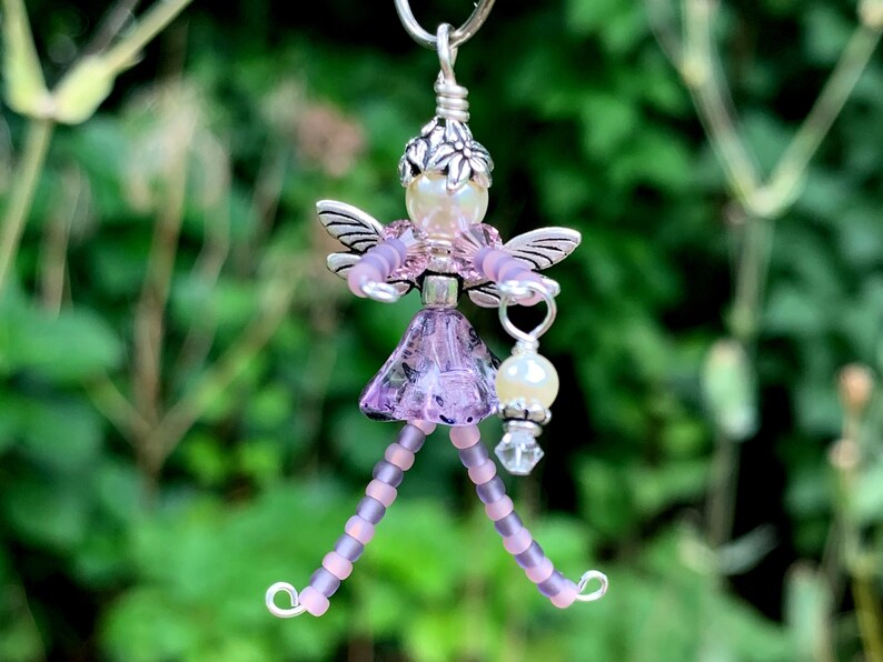 May include: A beaded fairy charm with a purple dress, wings, and a pearl and crystal embellishment. The fairy is holding a small lantern.