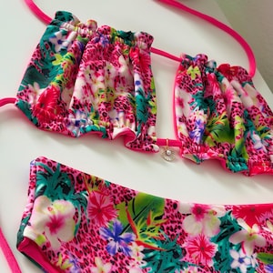 Peut inclure: Un maillot de bain deux pièces avec un imprimé floral et léopard vibrant. Le haut du bikini a des bords à volants et un dos nu. Le bas est taille haute. La palette de couleurs comprend le rose, le vert, le blanc et le bleu.