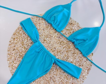 May include: A turquoise blue bikini set with a triangle top and a high-cut bottom. The bikini is on a white background with a round, textured surface.