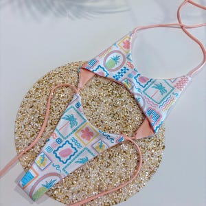 Puede incluir: Un conjunto de bikini de dos piezas con un estampado tropical. La parte superior e inferior del bikini presentan un fondo blanco con cuadrados coloridos, piñas, palmeras y flores. Los bordes y las correas son de color rosa claro.