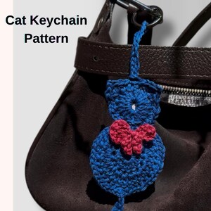 Peut inclure: Un porte-clés chat bleu au crochet avec un nœud rouge, accroché à un sac à main marron. Le porte-clés a un corps rond et une queue. Le texte "Cat Keychain Pattern" est visible.