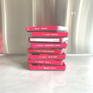May include: A stack of red Sizzix die-cutting machines with black foam inserts. Each machine is labeled with a different design, including "Elephant," "Filmstrip," "Gingerbread House," and "Telephone."