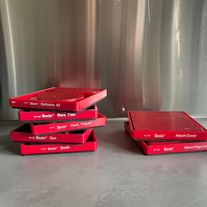 May include: A stack of red Sizzix die-cutting templates, including "Balloons #2", "Bare Tree", "Clock Filigree", "Sun", and "Scroll". Two additional templates are labeled "Album Cover" and "Album Page Inserts".