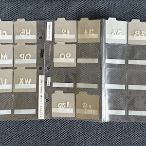 May include: A collection of craft stamps organized in clear plastic sleeves. The stamps are beige with white lettering and designs. The text "Sunshine Mini Shapes" is visible. The stamps are in various shapes and sizes, including letters, numbers, and shapes.