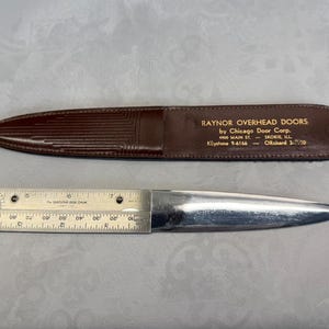May include: A vintage metal letter opener with a ruler, and a brown leather sheath. The sheath is embossed with the text "RAYNOR OVERHEAD DOORS by Chicago Door Corp."