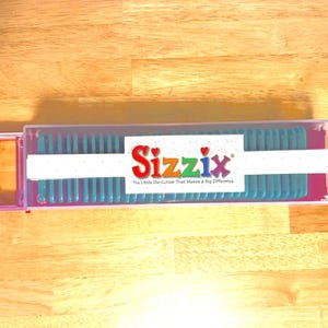 May include: A clear plastic Sizzix die-cutting storage case with a pink handle and accents. The case contains teal dies and a white label with the Sizzix logo and tagline. The case is on a wooden surface.