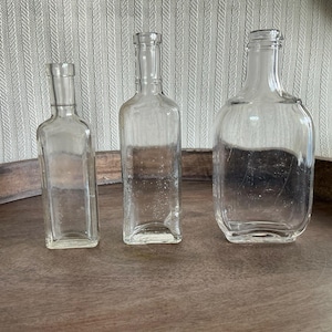 May include: Three clear glass bottles of varying shapes and sizes. The bottles are empty and sit on a wooden surface. The bottles are rectangular, square, and rounded. The background is a light-colored, textured fabric.