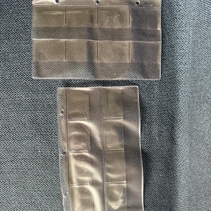 May include: Two clear plastic coin holder sheets. Each sheet has multiple rectangular pockets for storing coins. The sheets have three holes along the top edge for binder storage. The sheets are laying on a textured, dark blue surface.