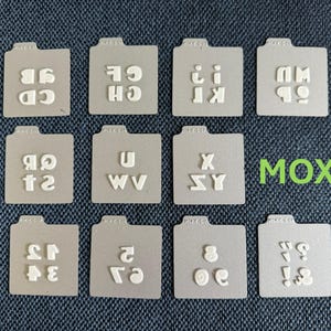 May include: A collection of gray, folder-shaped stamps with white letters and numbers. The word "MOXIE" is in green. The stamps include letters like "CD", "CH", "KI", "SY", and numbers like "34", "2", and symbols.