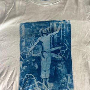 Puede incluir: Camiseta blanca con un estampado azul de una persona con los brazos levantados en una escena de bosque. El estampado tiene un estilo vintage y artístico. La camiseta tiene cuello redondo y una pequeña etiqueta azul con la palabra "DUNSH".