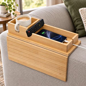 May include: A light-colored bamboo sofa arm tray with a flexible slatted design. It features a built-in square cup holder, a slot for a remote control, and a rectangular tray section holding a smartphone with a white charging cable attached.
