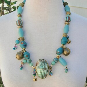 Könnte beinhalten: Eine türkisfarbene und goldene Perlenkette mit einem großen, ovalen, türkisfarbenen Anhänger. Die Kette enthält verschiedene Perlen, darunter kleine Glöckchen, und hat einen Boho-Stil.