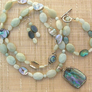 Puede incluir: Un conjunto de joyas que incluye un collar, una pulsera y pendientes. El conjunto presenta cuentas ovaladas de color verde p&aacute;lido, detalles iridiscentes de concha de abul&oacute;n y un cierre plateado. El collar tiene un colgante grande con una concha de abul&oacute;n.