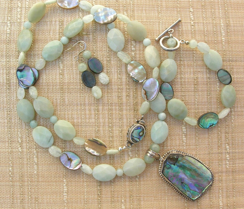 Puede incluir: Un conjunto de joyas que incluye un collar, una pulsera y pendientes. El conjunto presenta cuentas ovaladas de color verde p&aacute;lido, detalles iridiscentes de concha de abul&oacute;n y un cierre plateado. El collar tiene un colgante grande con una concha de abul&oacute;n.