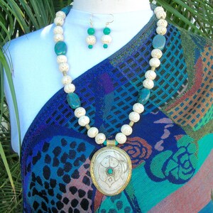 Può includere: Una collana di perline color crema con accenti turchesi e un paio di orecchini abbinati. La collana presenta un grande pendente ovale con una cornice color oro e una pietra centrale turchese. I gioielli sono esposti su un manichino bianco.