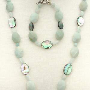 Puede incluir: Un conjunto de joyas de tres piezas con collar, pulsera y pendientes. El collar y la pulsera tienen cuentas ovaladas de color azul claro con detalles iridiscentes de concha de abul&oacute;n. El collar tiene un colgante grande de color verde oscuro. Los pendientes tienen colgantes ovalados negros.