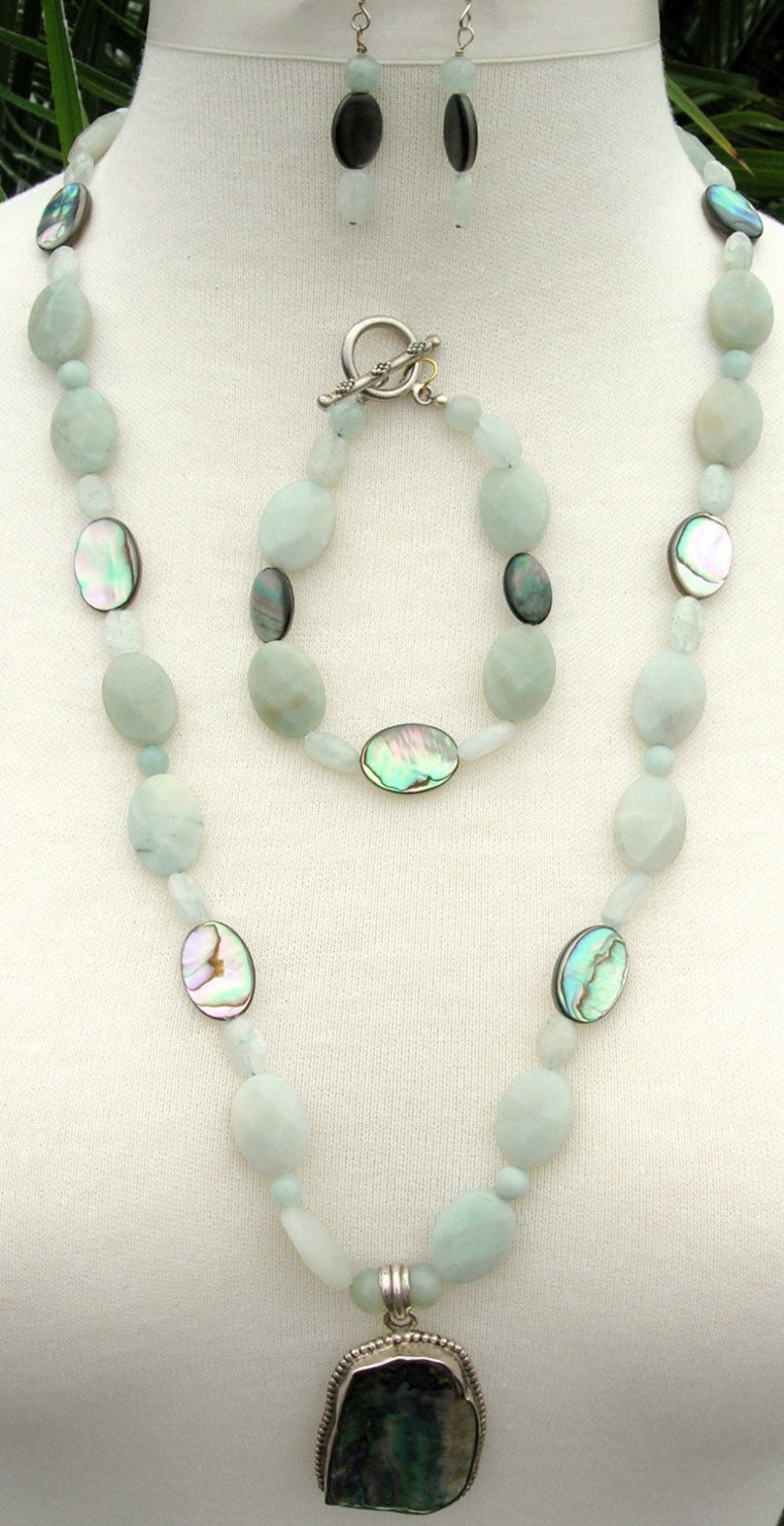 Puede incluir: Un conjunto de joyas de tres piezas con collar, pulsera y pendientes. El collar y la pulsera tienen cuentas ovaladas de color azul claro con detalles iridiscentes de concha de abul&oacute;n. El collar tiene un colgante grande de color verde oscuro. Los pendientes tienen colgantes ovalados negros.